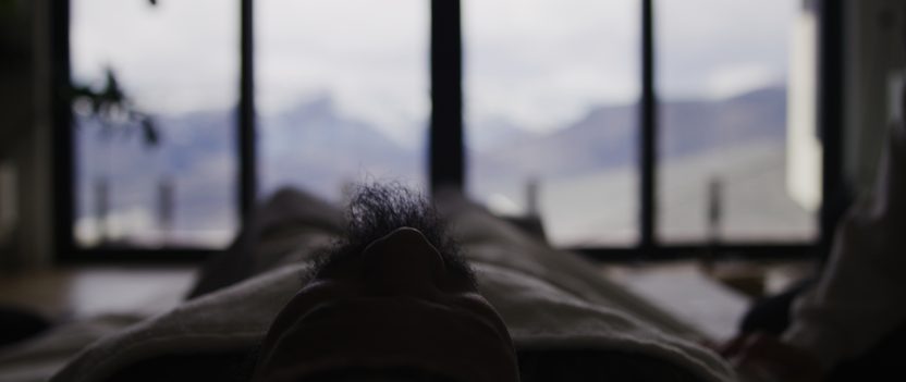 A figure lying beneath a blanket, silhouetted against a window and mountains beyond